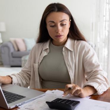 Mulher estudando contas financeiras em casa, com laptop, calculadora e papéis sobre a mesa, em um ambiente doméstico bem iluminado, ideal para gerenciamento financeiro.