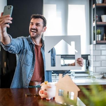 Homem feliz tirando selfie na cozinha moderna enquanto segura uma casa de papelão com uma decoração aconchegante e plantas ao redor.