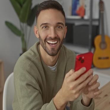 Homem sorridente segurando um smartphone vermelho em ambiente interno, com guitarra e plantas ao fundo, promovendo a palavra-chave 