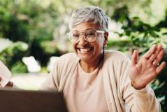 Mulher idosa sorrindo e acenando durante uma videochamada, transmitindo alegria e conexão emocional ao usar a tecnologia para manter contato com entes queridos.