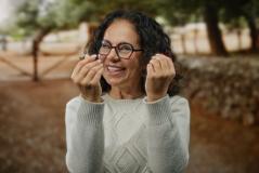 Mulher sorridente com óculos, vestindo suéter cinza, ao ar livre, expressando felicidade e alegria com um fundo de árvores e natureza.