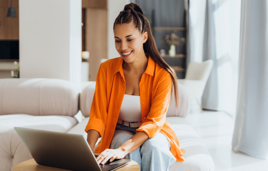 Uma mulher jovem e sorridente utiliza um notebook sobre uma mesa de centro enquanto está sentada em um sofá claro.
