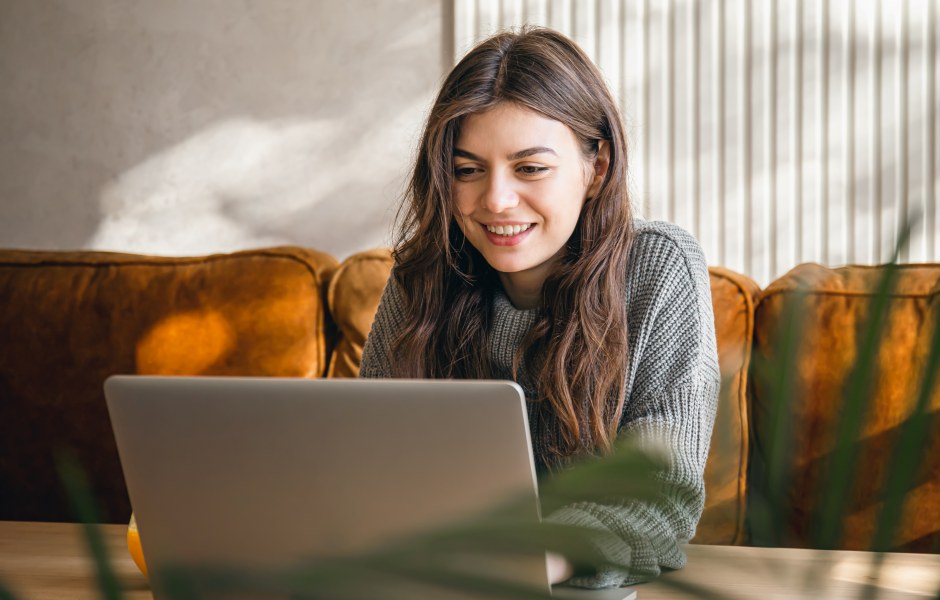 Jovem sorridente usa o laptop confortavelmente na sala de casa, com sofá mostarda e luz natural ao fundo, transmitindo bem-estar e praticidade no dia a dia.