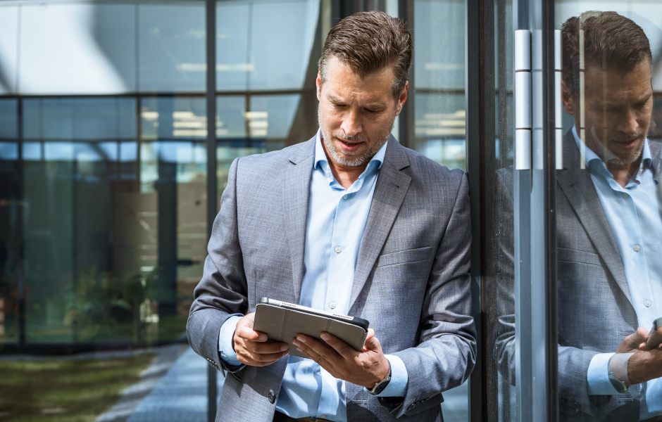 Homem de negócios usando tablet ao lado de vidro com reflexo, em ambiente corporativo moderno