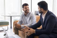 Dois profissionais em trajes sociais conversam de forma amigável em um escritório moderno, com um notebook sobre a mesa.