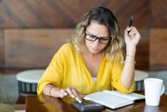Mulher de óculos analisa contas com expressão preocupada, usando calculadora e caderno sobre a mesa, com uma xícara de café ao lado.