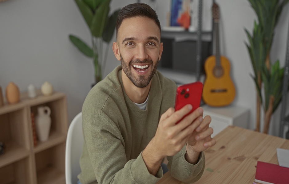 Homem sorridente segurando um smartphone vermelho em ambiente interno, com guitarra e plantas ao fundo, promovendo a palavra-chave 