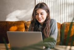 Jovem sorridente usa o laptop confortavelmente na sala de casa, com sofá mostarda e luz natural ao fundo, transmitindo bem-estar e praticidade no dia a dia.