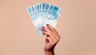 Mão segurando várias cédulas de dinheiro de 100 reais, destaque para a cor azul das notas e a expressão de valor e riqueza.