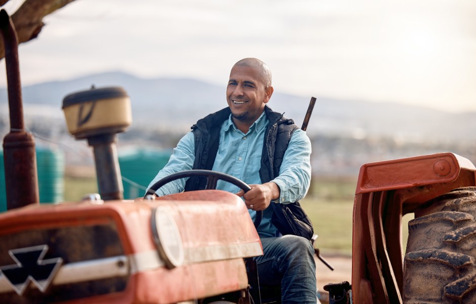 Agricultor sorridente conduz um trator vermelho no campo, com paisagem rural e montanhas ao fundo, transmitindo satisfação e orgulho no trabalho no campo.