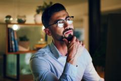 Um homem de barba e óculos olha pensativo para o lado enquanto apoia a mão no queixo em um escritório. Ele veste uma camisa social clara e está em um ambiente moderno com iluminação suave ao fundo.