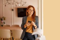 Mulher com cabelo vermelho, usando óculos e roupa profissional cinza blazer e blusa na cor mostarda, sorrindo e segurando um smartphone em ambiente moderno com decoração elegante
