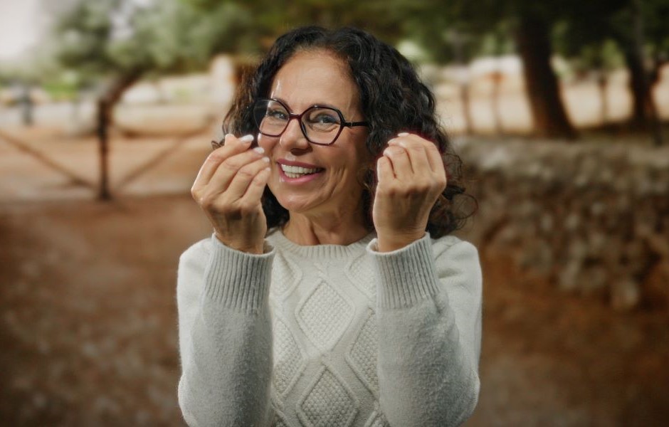 Mulher sorridente com óculos, vestindo suéter cinza, ao ar livre, expressando felicidade e alegria com um fundo de árvores e natureza.