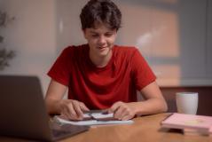 Jovem adolescente estudando em casa, utilizando computador e caderno, com expressão concentrada, ambiente acolhedor com luminária e objetos de estudo.