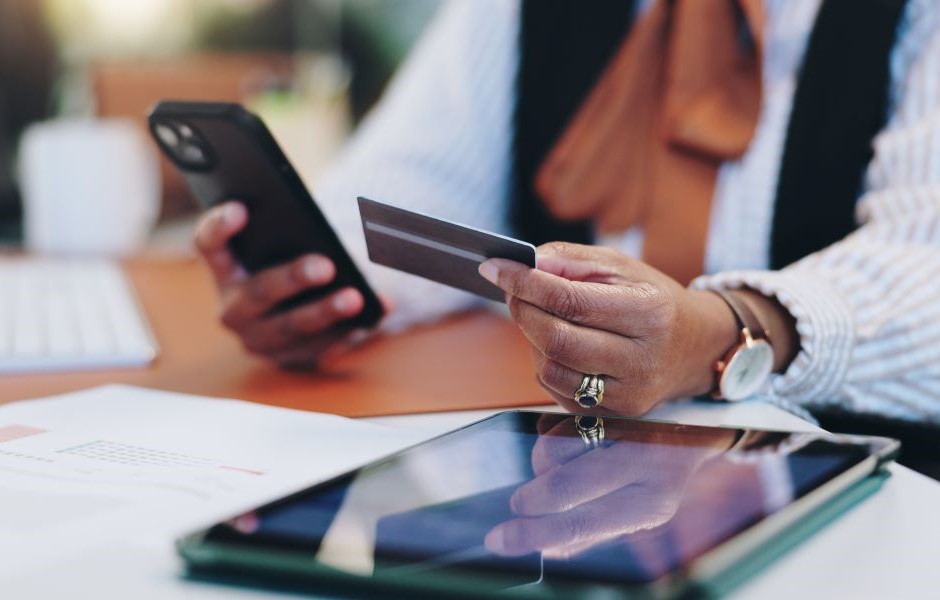 Pessoa usando um smartphone e um cartão de crédito em uma mesa de trabalho, com vários dispositivos eletrônicos ao redor, representando transações bancárias e compras online.