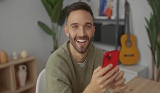 Homem sorridente segurando um smartphone vermelho em ambiente interno, com guitarra e plantas ao fundo, promovendo a palavra-chave 