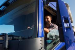 Caminhoneiro sorridente apoia o braço na janela aberta de sua cabine azul, com céu ensolarado ao fundo, transmitindo confiança e satisfação na profissão.