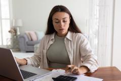 Mulher estudando contas financeiras em casa, com laptop, calculadora e papéis sobre a mesa, em um ambiente doméstico bem iluminado, ideal para gerenciamento financeiro.