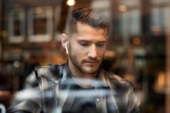 Homem jovem de cabelo curto e barba bem aparada, vestindo camisa xadrez escura. Ele está olhando para baixo com expressão séria e usa um fone de ouvido sem fio branco na orelha direita. A foto foi tirada através do vidro de uma janela, que reflete luzes desfocadas ao fundo.