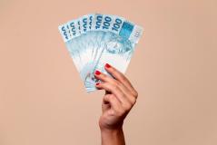 Mão segurando várias cédulas de dinheiro de 100 reais, destaque para a cor azul das notas e a expressão de valor e riqueza.