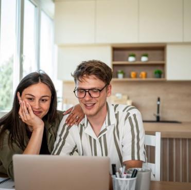 Casal sorridente assistindo a algo no laptop sentado na cozinha moderna com iluminação natural, elementos decorativos e ambiente acolhedor.