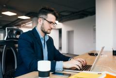 Jovem executivo de óculos digita concentrado no laptop em escritório moderno, com café e celular sobre a mesa, demonstrando foco e produtividade.