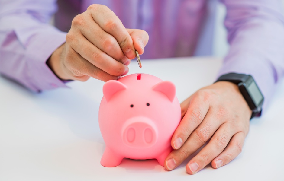 Mão depositando uma moeda em um cofrinho rosa, simbolizando o hábito de poupar e a importância do planejamento financeiro para o futuro.