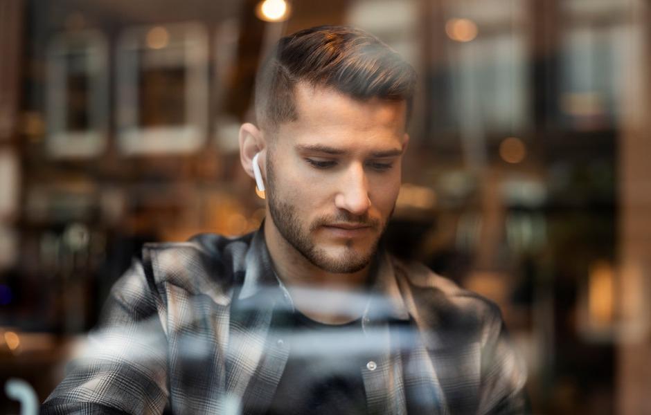 Homem jovem de cabelo curto e barba bem aparada, vestindo camisa xadrez escura. Ele está olhando para baixo com expressão séria e usa um fone de ouvido sem fio branco na orelha direita. A foto foi tirada através do vidro de uma janela, que reflete luzes desfocadas ao fundo.