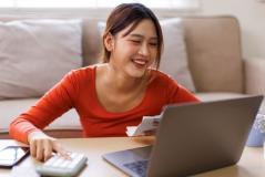 Mulher sorridente usando um laptop e segurando uma fatura, estudando ou realizando tarefas administrativas em casa, com expressão de satisfação.