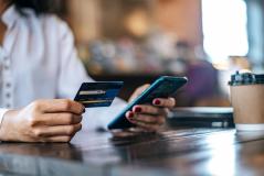 Mulher realiza pagamento pelo smartphone segurando cartão de crédito numa cafeteria, com copo de café descartável ao lado sobre a mesa.