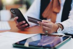 Pessoa usando um smartphone e um cartão de crédito em uma mesa de trabalho, com vários dispositivos eletrônicos ao redor, representando transações bancárias e compras online.