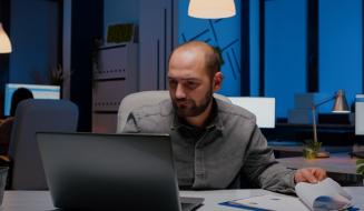 Profissional analisa relatórios e gráficos no laptop durante a madrugada em escritório com iluminação azulada, transmitindo dedicação intensa ao trabalho.
