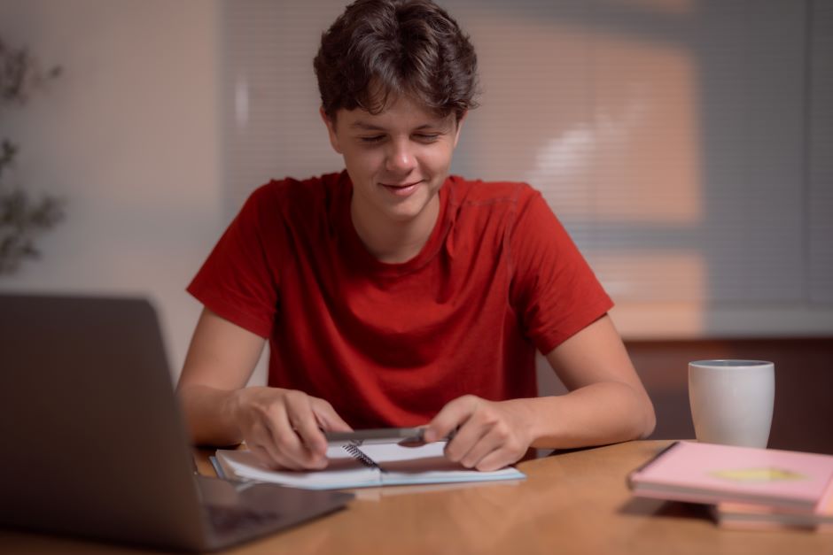 Jovem adolescente estudando em casa, utilizando computador e caderno, com expressão concentrada, ambiente acolhedor com luminária e objetos de estudo.
