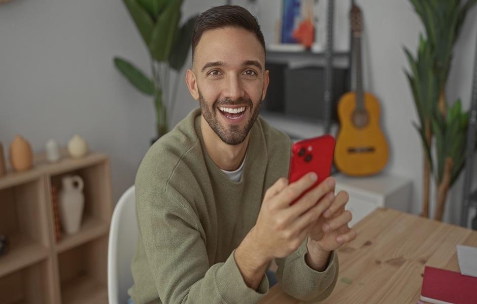 Um homem jovem e sorridente segura um celular vermelho enquanto está sentado à frente de uma mesa de madeira.
