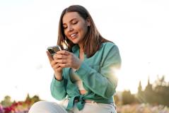 Jovem sorridente usa o smartphone ao ar livre num parque florido, com luz dourada do pôr do sol ao fundo criando uma atmosfera leve e descontraída.
