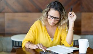 Mulher de óculos analisa contas com expressão preocupada, usando calculadora e caderno sobre a mesa, com uma xícara de café ao lado.