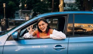 Mulher sorridente dentro de um carro azul, olhando pela janela e fazendo o sinal de 'ok' com a mão.