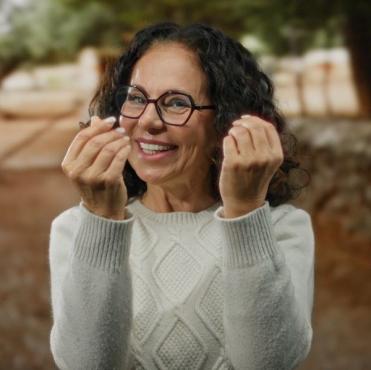 Mulher sorridente com óculos, vestindo suéter cinza, ao ar livre, expressando felicidade e alegria com um fundo de árvores e natureza.