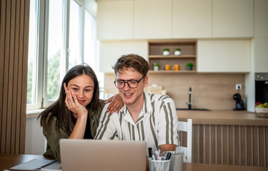 Casal sorridente assistindo a algo no laptop sentado na cozinha moderna com iluminação natural, elementos decorativos e ambiente acolhedor.