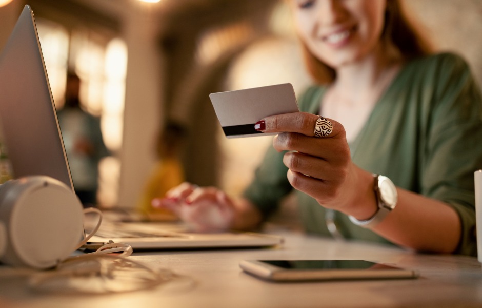 Mulher sorridente segura cartão de crédito enquanto realiza uma compra online no laptop, num ambiente com iluminação aconchegante e quente.