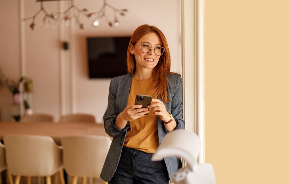 Mulher com cabelo vermelho, usando óculos e roupa profissional cinza blazer e blusa na cor mostarda, sorrindo e segurando um smartphone em ambiente moderno com decoração elegante