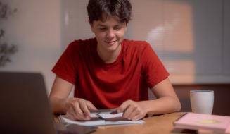 Jovem adolescente estudando em casa, utilizando computador e caderno, com expressão concentrada, ambiente acolhedor com luminária e objetos de estudo.
