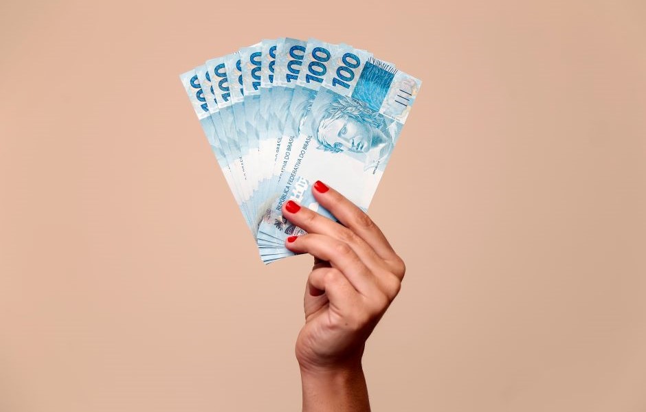 Mão segurando várias cédulas de dinheiro de 100 reais, destaque para a cor azul das notas e a expressão de valor e riqueza.