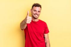 Homem sorridente de camiseta vermelha faz um sinal de positivo com o polegar em destaque. Ele está posicionado à frente de um fundo amarelo vibrante, exibindo uma tatuagem no braço.