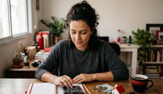 De cabelos cacheados presos, jovem utiliza uma calculadora sobre um caderno aberto para planejar o orçamento doméstico. Na mesa de madeira, moedas, cédulas de dinheiro, uma miniatura de casa com telhado vermelho e uma caneca de café completam o cenário de finanças pessoais.