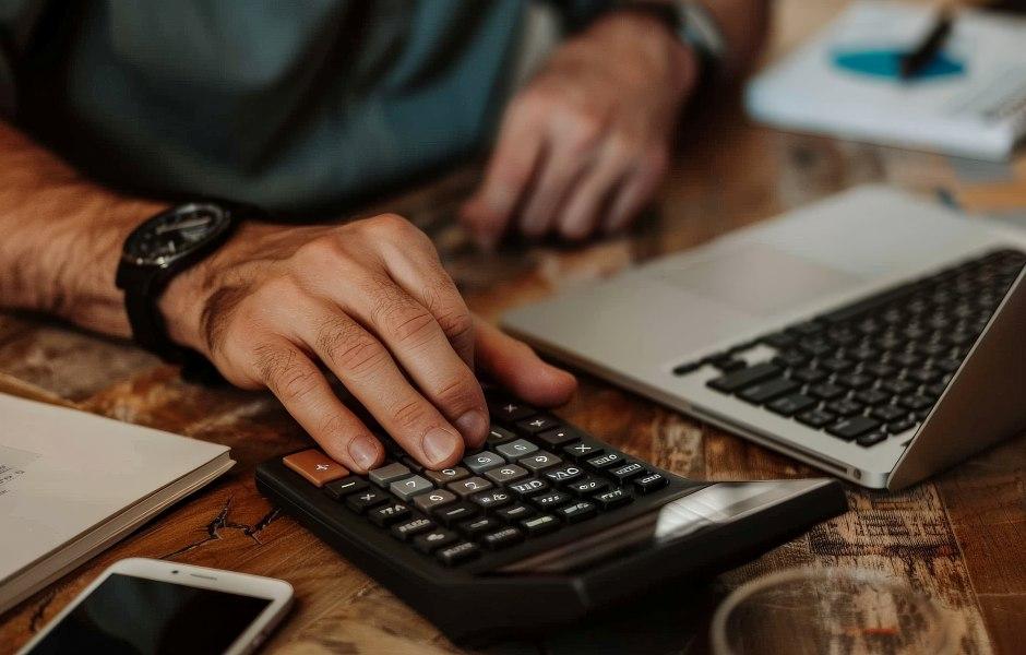 Uma mão masculina com um relógio preto utiliza uma calculadora sobre uma mesa de madeira rústica.