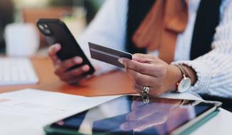 Pessoa usando um smartphone e um cartão de crédito em uma mesa de trabalho, com vários dispositivos eletrônicos ao redor, representando transações bancárias e compras online.