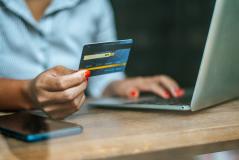 Close de mulher segurando cartão de crédito azul enquanto digita no laptop, realizando um pagamento online com celular ao lado sobre a mesa.