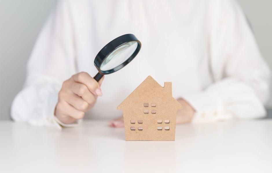 Uma mão segura uma lupa apontada para uma pequena miniatura de casa feita de papelão ou madeira clara.