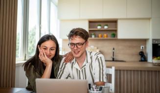 Casal sorridente assistindo a algo no laptop sentado na cozinha moderna com iluminação natural, elementos decorativos e ambiente acolhedor.